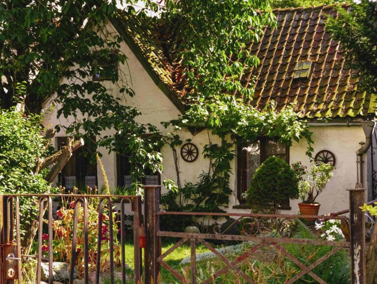 A rustic white cottage with a weathered tiled roof surrounded by lush green trees, plants, climbing vines, decorative wooden wheels, and a rusty iron gate in front.