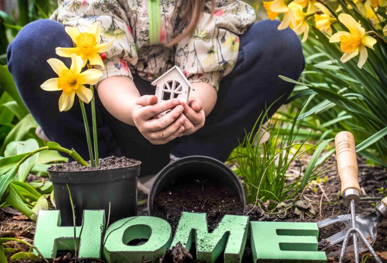 A child squatting in a garden holding a small wooden miniature house in their hands, surrounded by blooming yellow daffodils, potted soil, gardening tools, and wooden letters spelling "HOME".